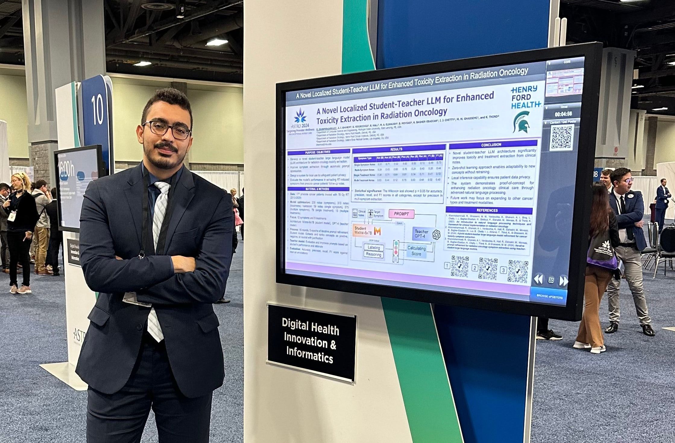 Reza with arms crossed next to a digital poster screen titled “A Novel Localized Student-Teacher LLM for Enhanced Toxicity Extraction in Radiation Oncology”.