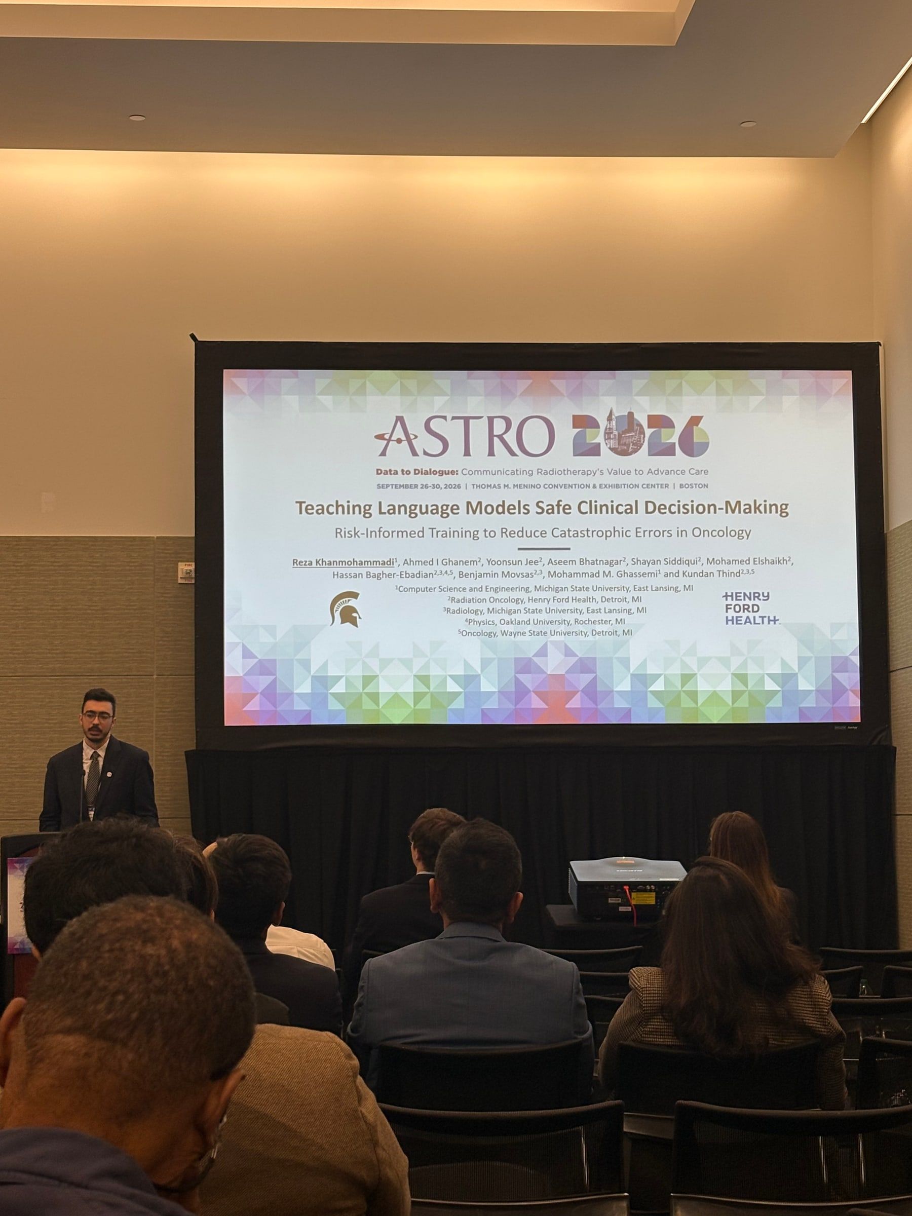 Reza at a podium beside a large screen showing the title slide “Teaching Language Models Safe Clinical Decision-Making”, with the audience seen from behind.